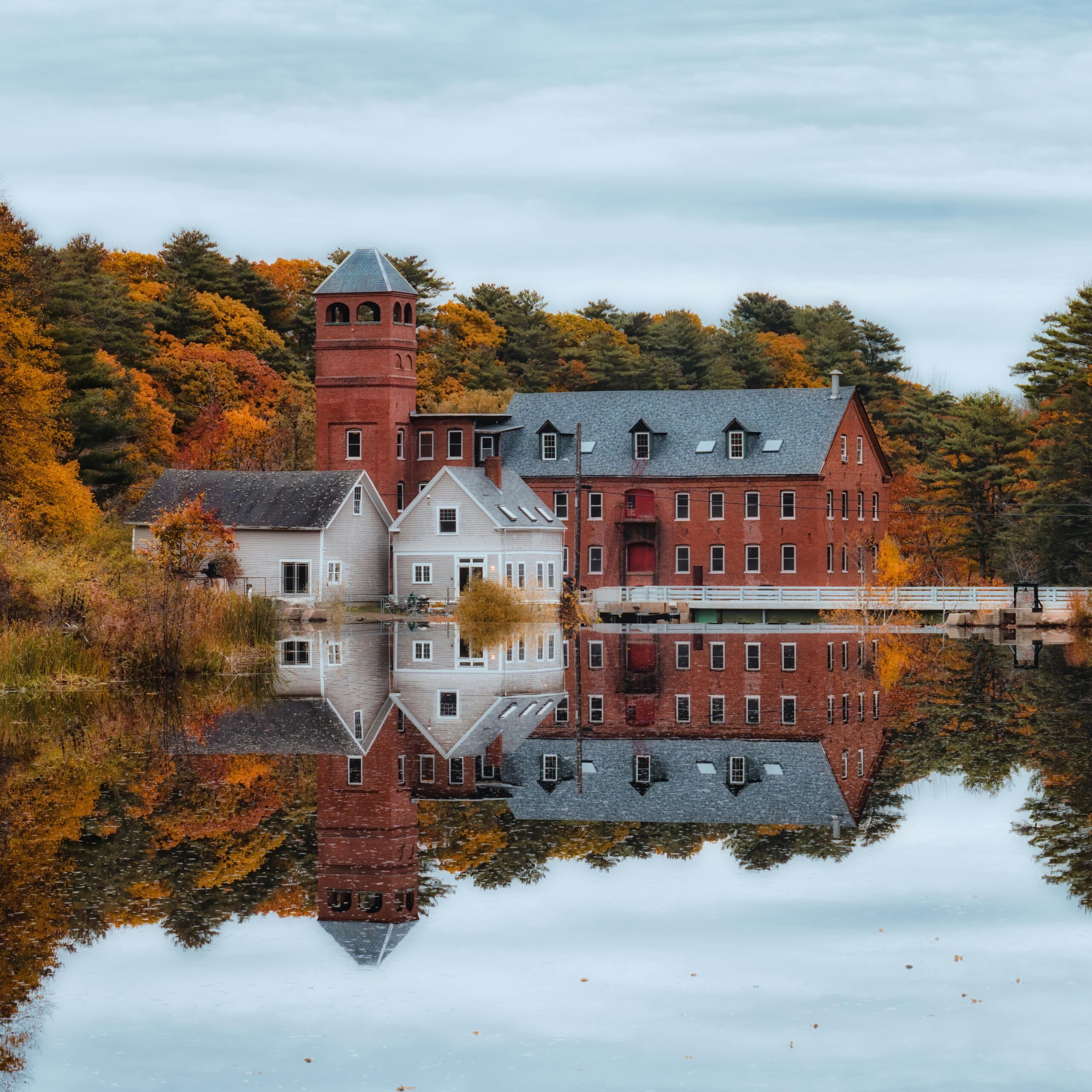Old manor with brick tower located on shore of calm pond surrounded by autumn trees under cloudy sky